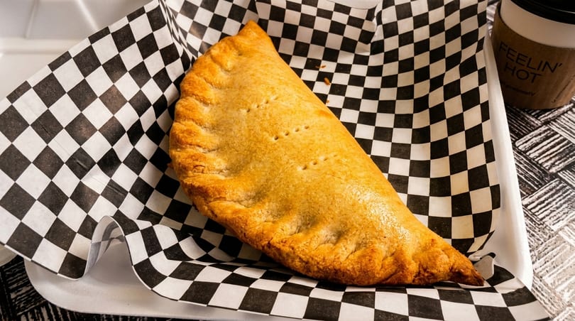 Golden-brown calzone with crimped edges, served on black and white checkered paper with a coffee cup.
