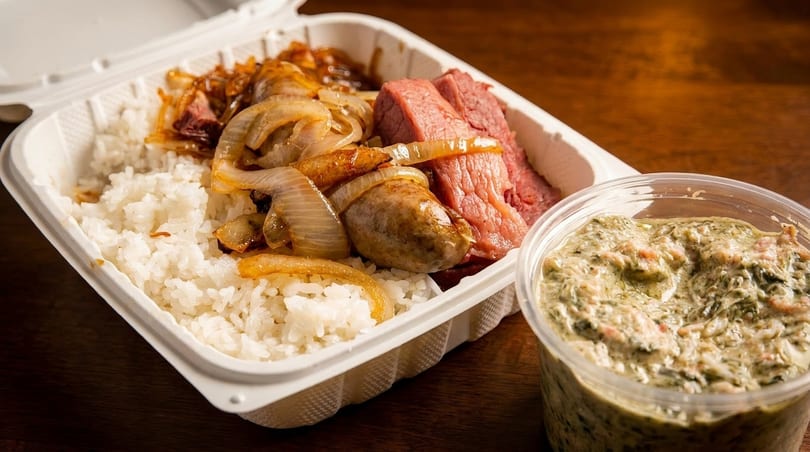 Takeout container with white rice, sliced meat, sausage, caramelized onions, and a side of green dip.