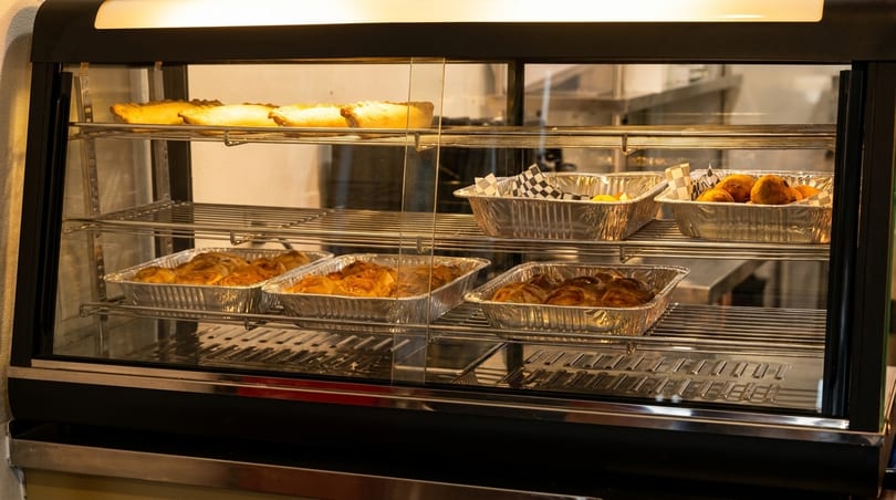 A brightly lit display case showcases various baked goods and pastries in foil containers.