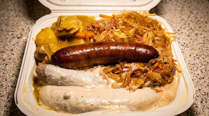 Takeout container with two sausages, curry chicken, potatoes, and glass noodles with vegetables.