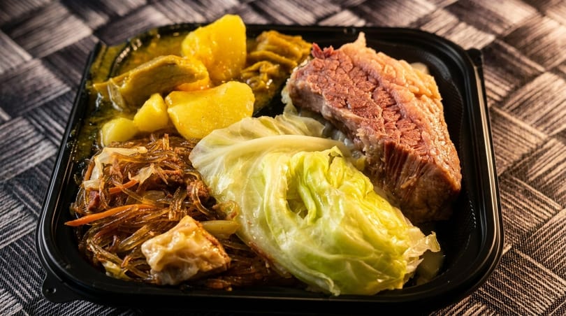 A black takeout container filled with corned beef, cabbage, noodles, and potatoes in sauce.