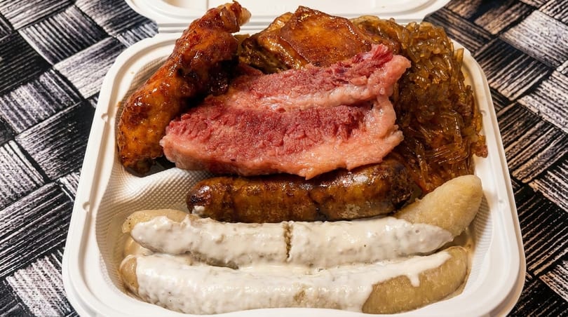 A takeout container filled with various meats, noodles, and plantains with white sauce.